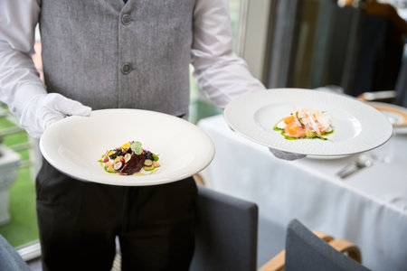 Cropped photo of male waiter in uniform carrying two plates of salads in a restaurantの写真素材