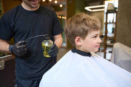Smiling barber spraying cologne water at little boy new stylish haircutの写真素材