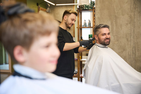 Caring father smiling watching qualified barber cutting his son hairの写真素材