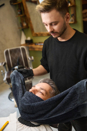 Focused barber carefully wrapping hot towel around his faceの写真素材