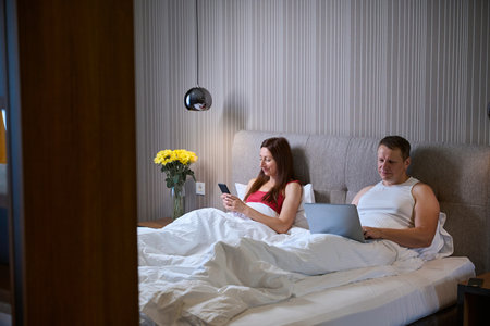 Married couple in bed with modern gadgetsの写真素材