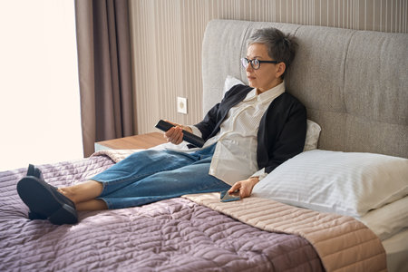 Caucasian grey-haired lady using tv remote control lying on bed in hotel roomの写真素材