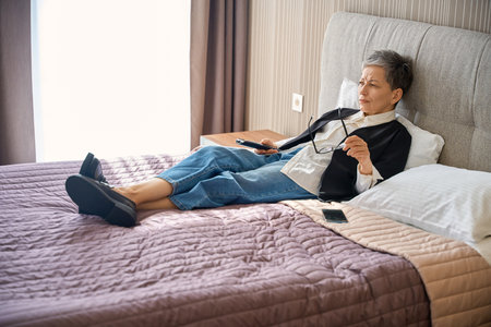 Serious adult woman watching tv while using remote control in hotel roomの写真素材