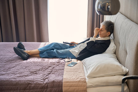 Tired adult woman holding remote control while watching tv in hotel roomの写真素材
