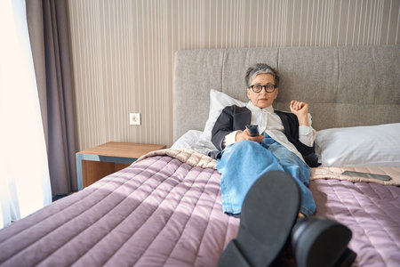 Caucasian senior lady switching with remote control while resting on bed in hotel roomの写真素材