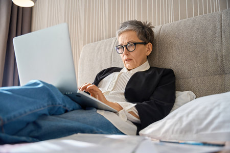 Senior businesswoman in glasses working with laptop sitting on bed in hotel roomの写真素材