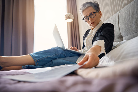 Adult businesswoman in glasses taking papers while working with laptop in hotel roomの写真素材