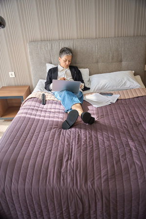 Tired adult businesswoman sleeping while sitting on bed with laptop in hotelの写真素材