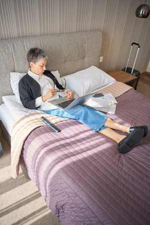 Adult businesswoman sleeping while holding glasses and laptop on legs in hotel roomの写真素材
