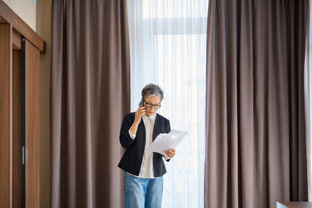 Serious adult businesswoman holding report while talking on the phone in hotelの写真素材