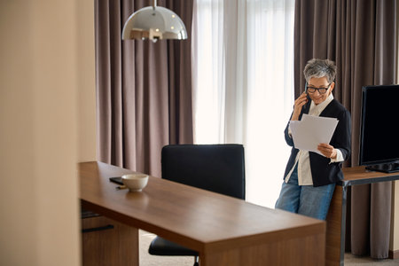 Caucasian senior woman reading documents while talking on the mobile phone in hotelの写真素材