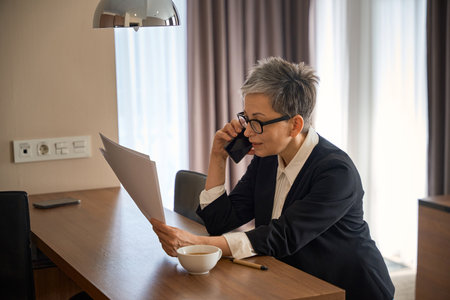 Adult businesswoman in glasses talking on the smartphone holding document in hotel roomの写真素材