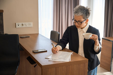 Senior lady filling out the form, holding cup of tea in hotel roomの写真素材