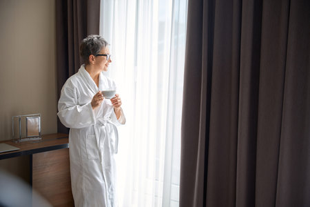 Happy senior woman in white bathrobe holding cup of tea, looking out the window in hotelの写真素材