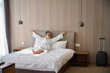Happy mature woman wearing bathrobe sitting on the bed holding hands to the side in hotel roomの写真素材