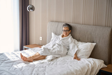 Happy adult woman in bathrobe sitting on the bed enjoying vacation in hotelの写真素材
