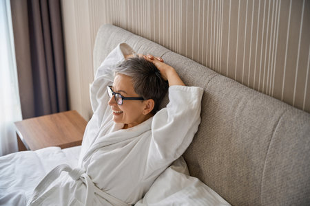 Smiling grey-haired woman wearing white bathrobe resting in hotel roomの写真素材