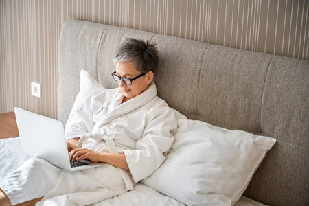 Smiling adult woman in white bathrobe typing on laptop sitting on bed in hotelの写真素材