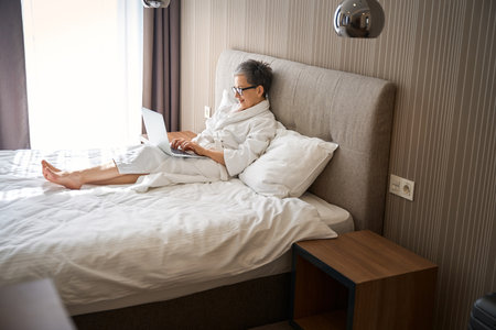 Adult woman in glasses wearing white bathrobe working with laptop in hotel roomの写真素材