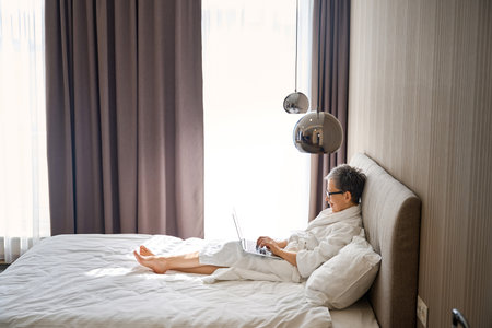 Mature woman wearing white bathrobe typing on laptop in hotel room, sitting on bedの写真素材