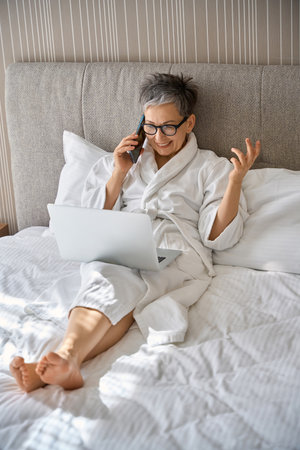 Pretty adult woman talking on mobile phone while working with laptop sitting on bed in hotelの写真素材
