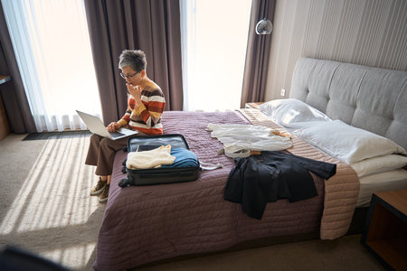 Senior woman sitting on the bed in hotel room while looking at laptop screenの写真素材