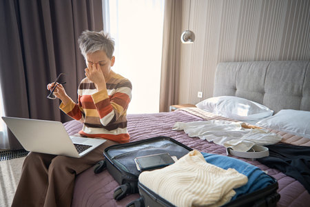 Tired senior businesswoman using laptop sitting on the bed in hotel roomの写真素材
