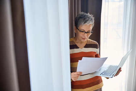 Smiling mature woman looking at document holding laptop in hotel roomの写真素材