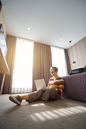 Happy senior lady sitting on floor while working with laptop in hotel roomの写真素材