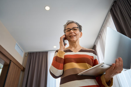 Happy businesswoman talking on the phone while holding laptop in hotel roomの写真素材