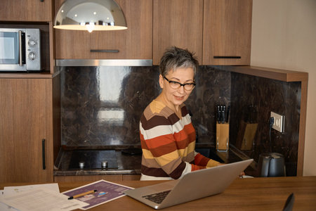 Senior woman making tea while looking at laptop screen in hotel roomの写真素材