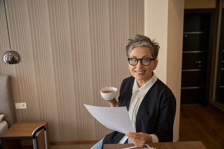 Smiling senior businesswoman in glasses drinking tea and holding papers in hotel roomの写真素材