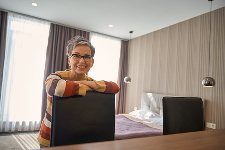 Smiling adult lady in glasses leaning on a chair posing at camera in hotel roomの写真素材