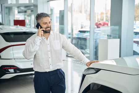 Happy man standing near new car and talking on mobile phone in auto showroomの写真素材