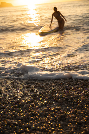 Adult woman carrying paddle board at ocean beach during sunset