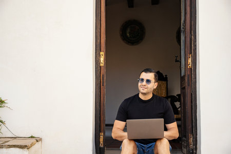 Smiling adult man holding laptop on legs, sitting on the street near entrance at home in Guadalest, Spainの写真素材