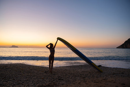 Young woman holding paddleboard on serene beach at sunsetの写真素材