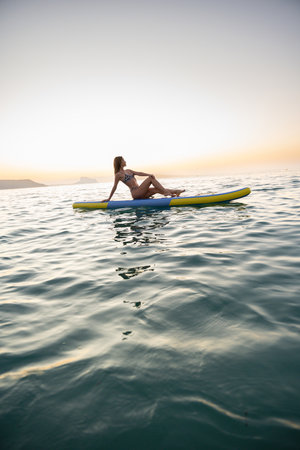 Young woman sitting on paddleboard in ocean at sunsetの写真素材