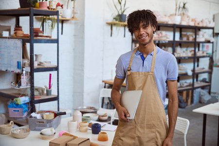 Young man smiling while holding laptop in pottery workshop