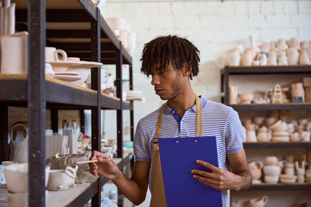 Focused adult man examining pottery in workshop with clipboardの写真素材
