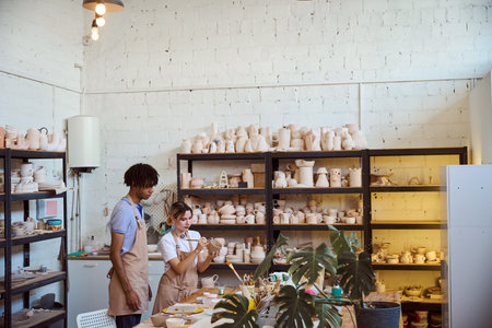 Young woman painting ceramic object in pottery studio with man observing