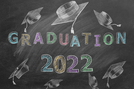 Hand drawn text GRADUATION 2022 and graduation caps on blackboard.