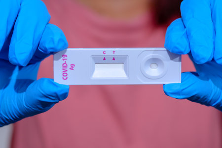 Woman wearing blue medical gloves and holding COVID-19 (SARS-CoV-2) First Antigen Test kit device to help in the Rapid Detection testing at home. Coronavirus global pandemic outbreak crisis âconcept.の写真素材