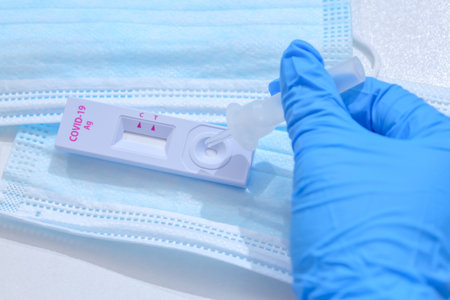 Woman wearing blue medical gloves and holding COVID-19 (SARS-CoV-2) First Antigen Test kit device to...