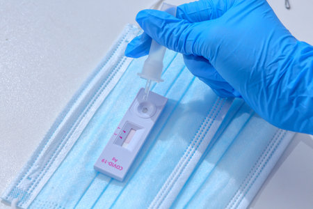 Woman wearing blue medical gloves and holding COVID-19 (SARS-CoV-2) First Antigen Test kit device to...