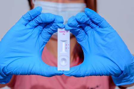 Woman wearing blue medical gloves and holding COVID-19 (SARS-CoV-2) First Antigen Test kit device to help in the Rapid Detection testing at home. Coronavirus global pandemic outbreak crisis âconcept.の写真素材