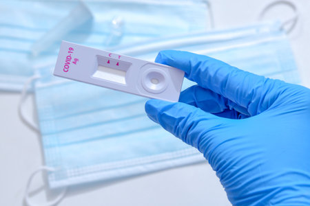 Woman wearing blue medical gloves and holding COVID-19 (SARS-CoV-2) First Antigen Test kit device to help in the Rapid Detection testing at home. Coronavirus global pandemic outbreak crisis âconcept.の写真素材