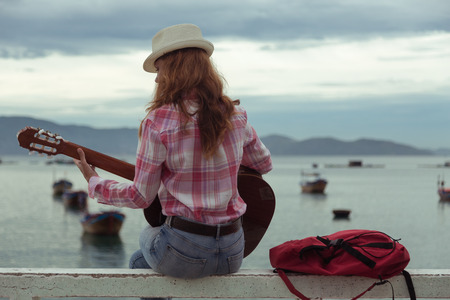 beautiful red-haired girl sitting on a fence with a guitar on the beachの写真素材