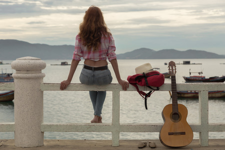 beautiful red-haired girl sitting on a fence with a guitar on the beachの写真素材