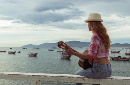 beautiful red-haired girl sitting on a fence with a guitar on the beachの写真素材
